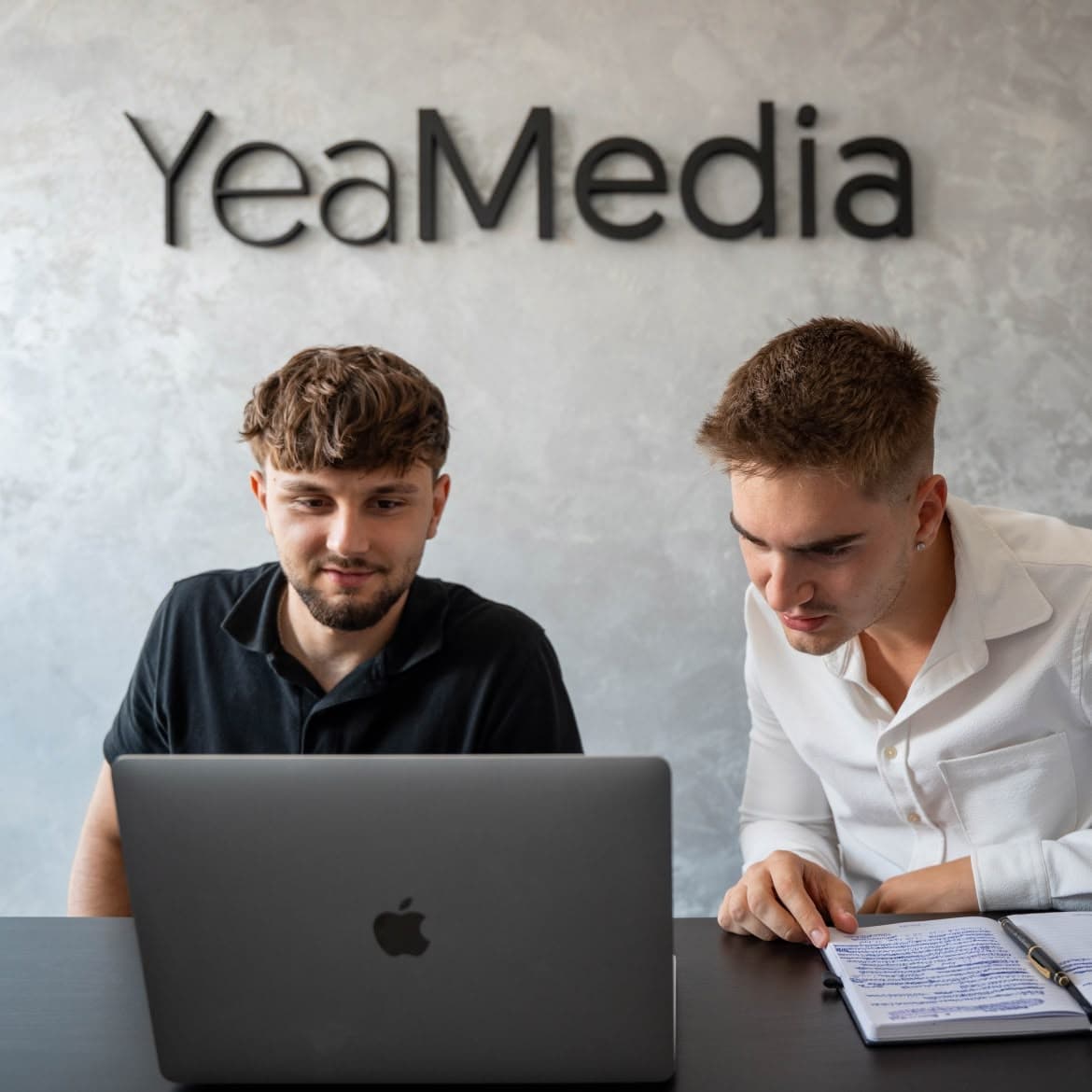 Zwei Mitarbeiter von YeaMedia arbeiten am Laptop vor dem Schriftzug an der Bürowand