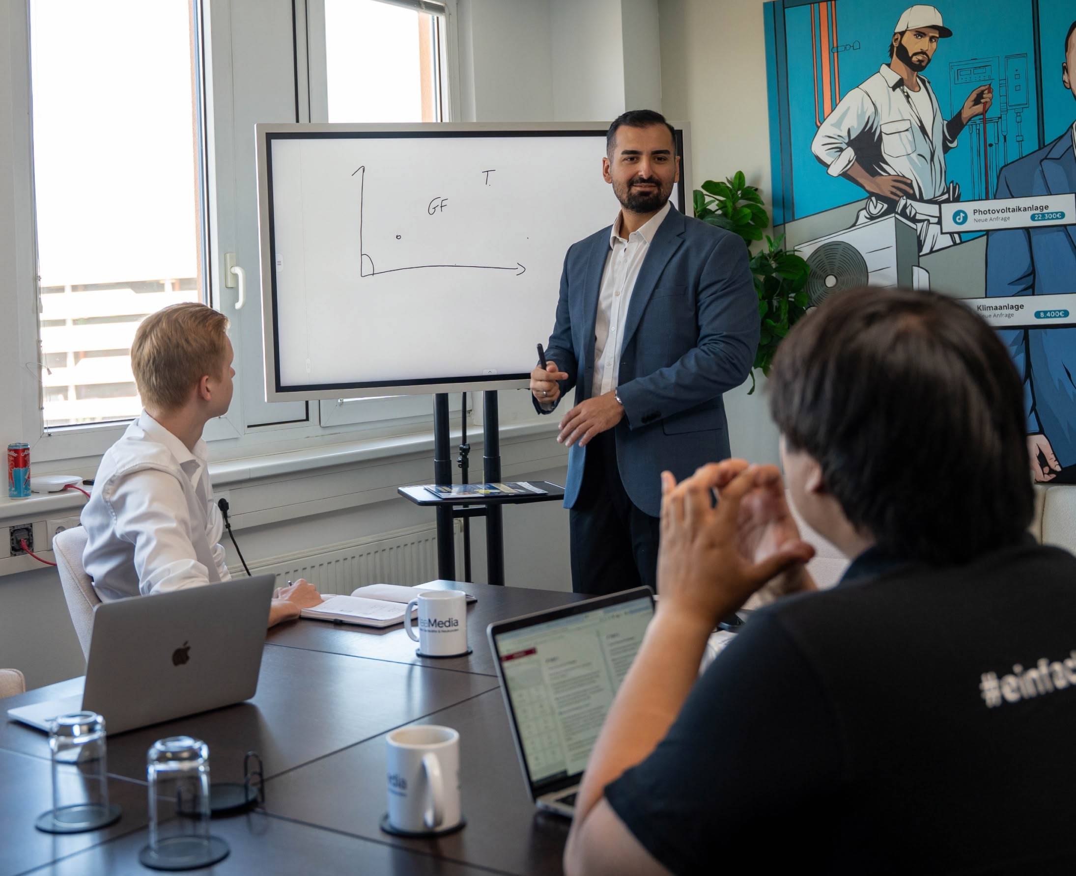 Yunus Akkaya erklärt zwei Mitarbeitern im Besprechungsraum etwas am Whiteboard