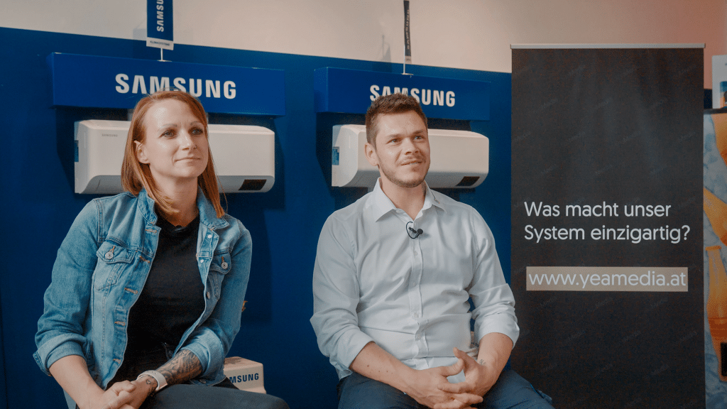 zwei Personen sitzen vor einer Wand wo Klimaanlagen von Samsung ausgestellt werden, sie hören einer Person die nicht im Bild ist zu